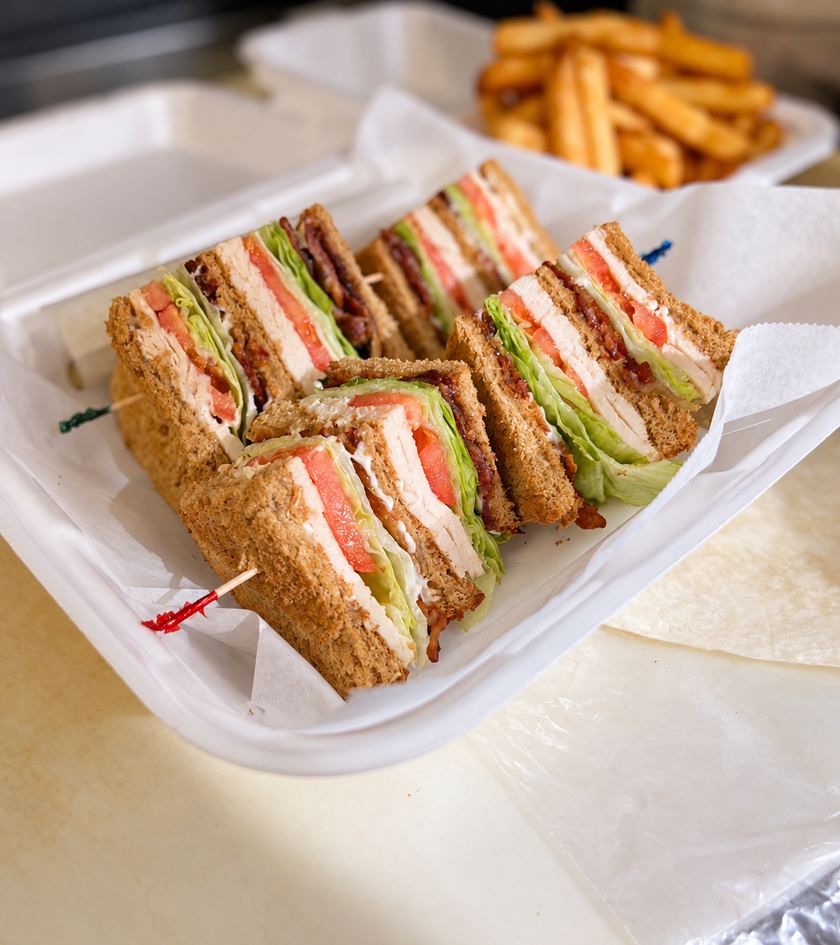Triple-decker club sandwich cut into quarters in a takeout container, with fries